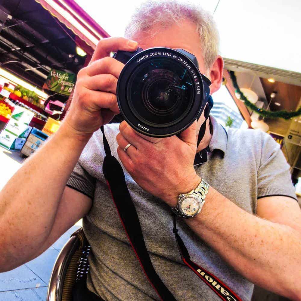 A Person Wearing A Gray Shirt And A Wristwatch, Resembling Alistair Pryde, Holds A Canon Camera, Capturing The Vibrant Scene At An Outdoor Market Or Café.