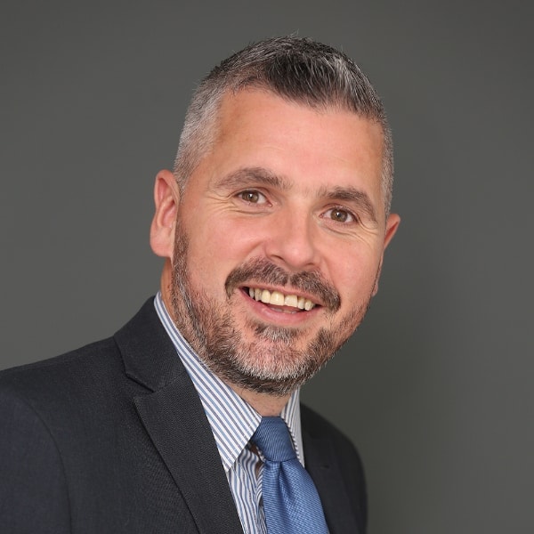 Chris Duffy, With Short, Graying Hair And A Beard, Is Dressed In A Dark Suit, Light Striped Shirt, And Blue Tie. He Smiles Warmly Against A Plain Gray Background.