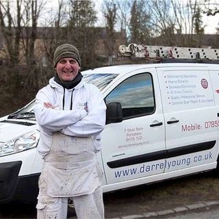 Darrel Young, Dressed In White Work Clothes And A Beanie, Stands With Arms Crossed In Front Of A White Van Featuring His Business Signage. The Van, Topped With A Ladder On Its Roof, Is Parked On A Road Surrounded By A Wooded Area.