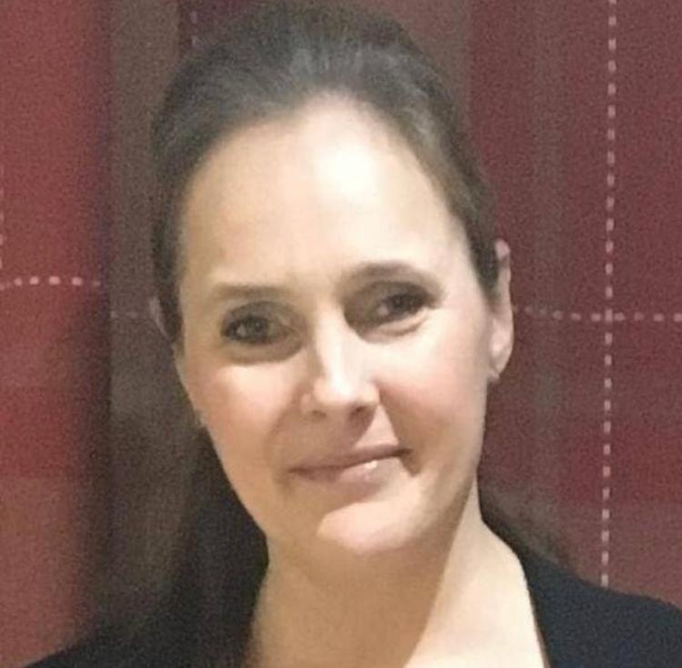 Kelly Combe, A Woman With Light Skin And Brown Hair Pulled Back, Is Smiling Gently And Looking Toward The Camera. She Is Standing In Front Of A Red Curtain With White Stitching.