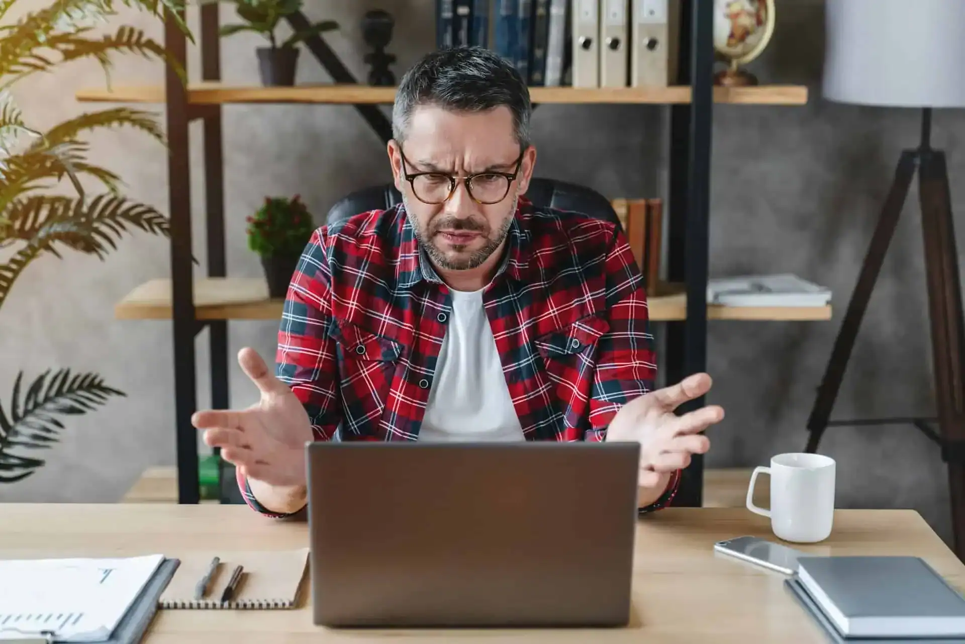 A man wearing glasses and a red plaid shirt sits at a desk, looking at his laptop with a frustrated expression and gesturing with his hands.