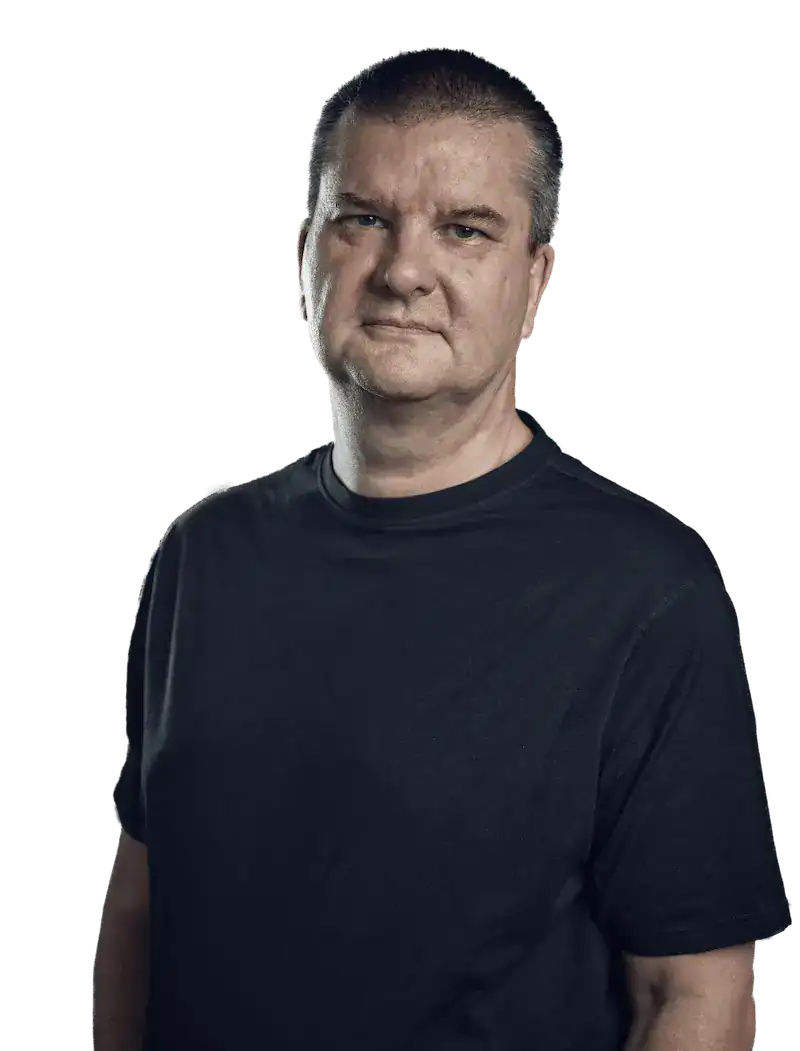 A middle-aged man with short hair wearing a plain black t-shirt stands facing the camera against a plain background.