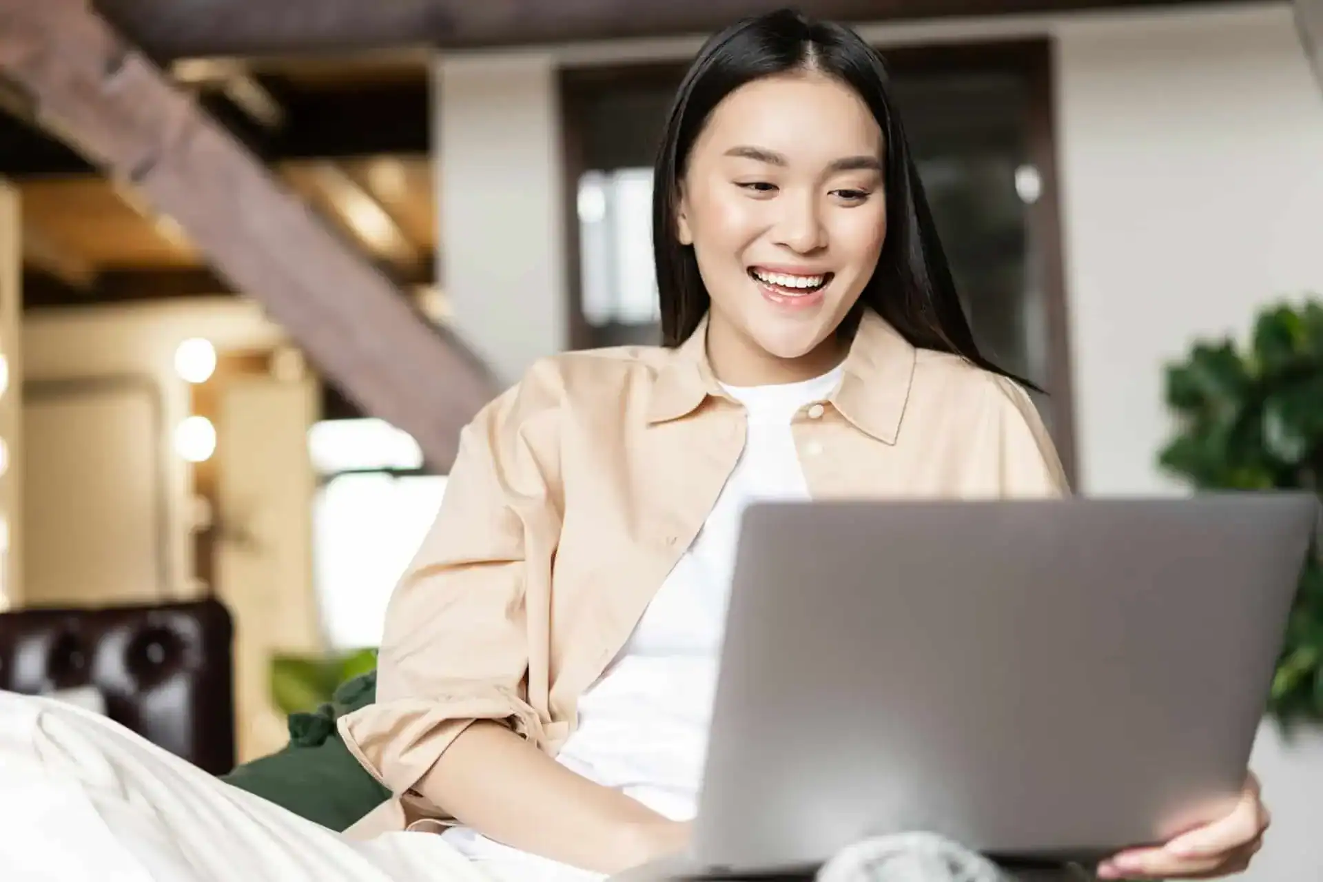 A woman sits indoors on a couch, smiling while looking at an open laptop on her lap.