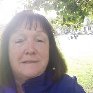 Morag Montgomery, A Woman With Shoulder Length Dark Hair, Stands Outdoors In A Park With Trees And Grass In The Background, Wearing A Purple Jacket.